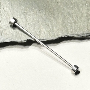 Op de afbeelding: Een zilverkleurige stropdas met een recht, dun ontwerp. De stropdas heeft aan elke kant een cilindrisch uiteinde. De achtergrond is een grijs, getextureerd oppervlak.