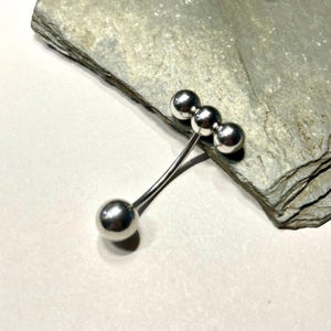 Puede incluir: Broche plateado de dise&ntilde;o abstracto con cuatro esferas. Tres esferas se agrupan en un extremo de un tallo curvo, y una esfera &uacute;nica en el otro. Se muestra sobre una superficie gris texturizada.