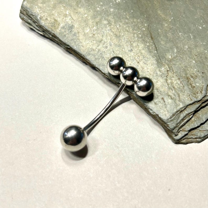 Puede incluir: Broche plateado de dise&ntilde;o abstracto con cuatro esferas. Tres esferas se agrupan en un extremo de un tallo curvo, y una esfera &uacute;nica en el otro. Se muestra sobre una superficie gris texturizada.