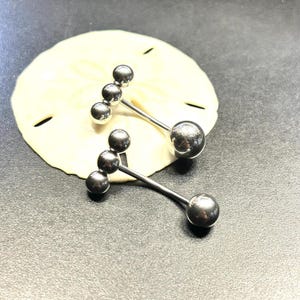 Puede incluir: Dos piercings para el ombligo de color plateado. Cada uno tiene una bola grande en la parte inferior y tres bolas m&aacute;s peque&ntilde;as en la parte superior, conectadas por una barra recta. Los anillos se muestran sobre una superficie clara y texturizada.
