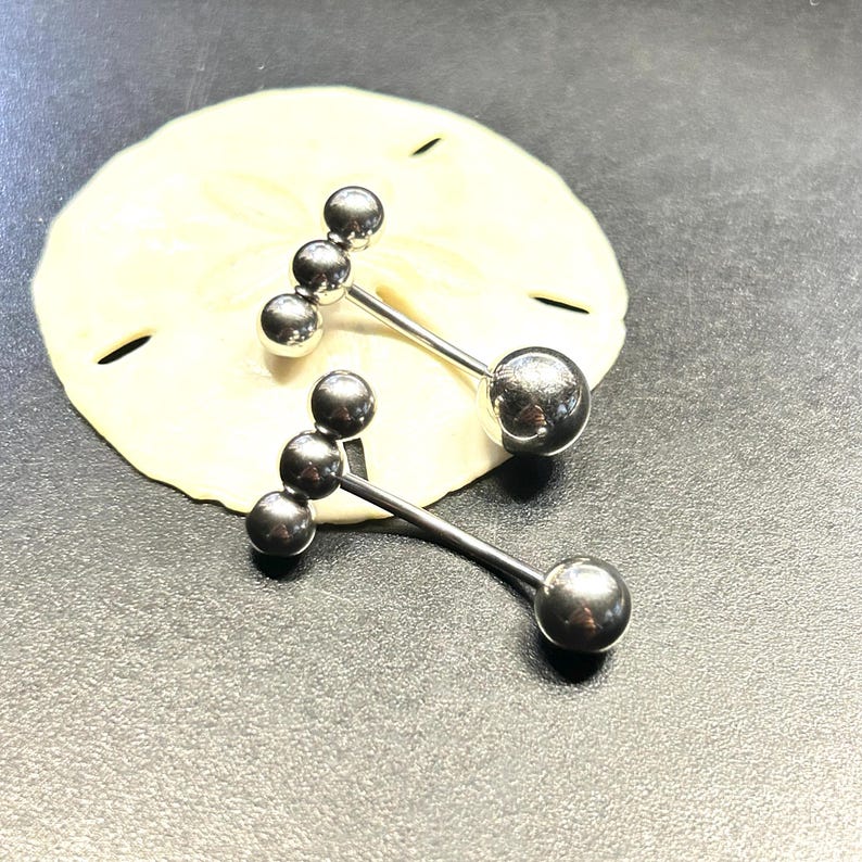 Puede incluir: Dos piercings para el ombligo de color plateado. Cada uno tiene una bola grande en la parte inferior y tres bolas m&aacute;s peque&ntilde;as en la parte superior, conectadas por una barra recta. Los anillos se muestran sobre una superficie clara y texturizada.