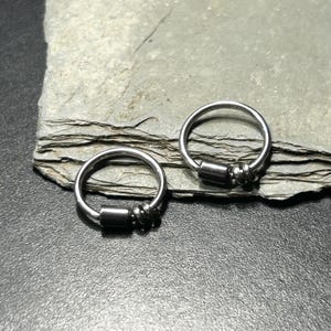 May include: Two silver-toned circular hoop rings, each featuring a smooth, dark metallic cylindrical bead and a smaller, textured coiled bead threaded onto the hoop. The rings are constructed from a thin, polished metal wire material.