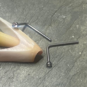 May include: Two silver nose rings with clear gem stones. The rings are bent at a 90 degree angle.