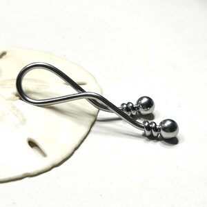 May include: A silver-toned metal body jewelry piece featuring a curved, twisted bar design that forms a loop at one end. The two ends of the bar are finished with polished spherical beads, each accented by a small, decorative grooved ring detail.
