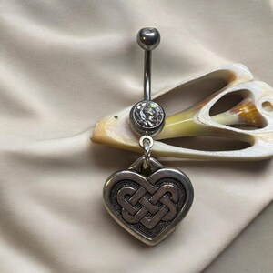 Könnte beinhalten: Silberner Bauchnabelring mit einem klaren Stein und einem baumelnden Herz-Anhänger mit einem keltischen Knotendesign.