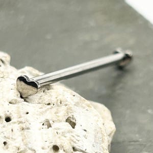 May include: A silver-toned industrial barbell body jewelry piece featuring a smooth, polished metal finish. The barbell has a small, solid heart-shaped end piece on one side and a rounded end on the other, designed for a piercing.