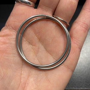 May include: A pair of silver hoop earrings, one slightly larger than the other, are shown in a hand.