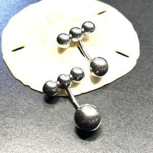 Puede incluir: Dos piercings para el ombligo de color plateado. Cada uno tiene una bola grande y redonda en la parte inferior y tres bolas m&aacute;s peque&ntilde;as en la parte superior, conectadas por una barra curva. Los anillos se muestran sobre una superficie blanca texturizada.