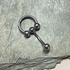 May include: A silver-colored body piercing jewelry piece. The jewelry features a curved barbell with three small, round balls and a straight barbell with a single ball at the end. The piece is made of a shiny, metallic material.