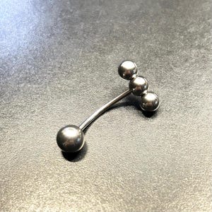 Puede incluir: Un piercing corporal de barra curva de color plateado con cuatro bolas esf&eacute;ricas. La barra est&aacute; hecha de metal brillante y se encuentra sobre un fondo oscuro.