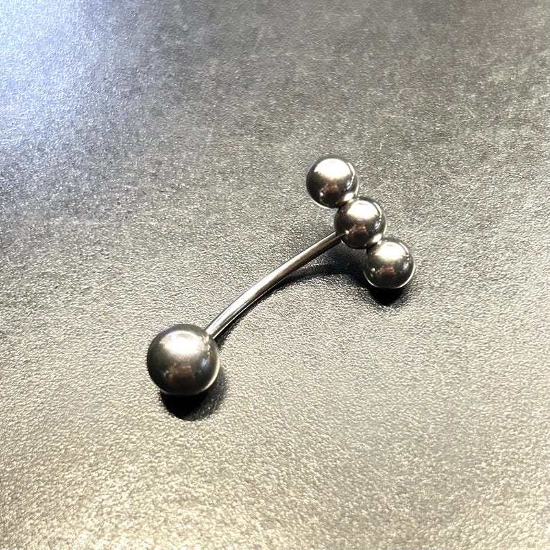 Puede incluir: Un piercing corporal de barra curva de color plateado con cuatro bolas esf&eacute;ricas. La barra est&aacute; hecha de metal brillante y se encuentra sobre un fondo oscuro.