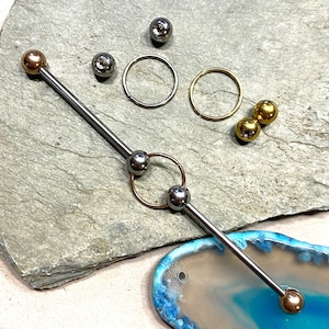Pode incluir: Uma barra de barbell prateada com duas bolas prateadas e um anel dourado no meio. A barra está sobre uma superfície de pedra cinza com uma pedra de ágata azul e branca no fundo.