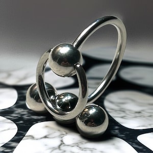 May include: A shiny, silver-colored metal ring with three small spheres and two larger spheres. The design is a combination of curved lines and spherical shapes. The background is a black and white pattern.