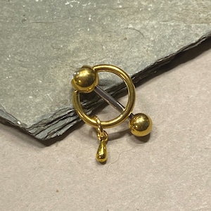 Könnte beinhalten: Ein goldfarbener Brustwarzenring mit kreisförmigem Design und einem hängenden Tropfenanhänger. Der Ring hat einen Langhantelstil mit einer Kugel an jedem Ende und einem kleinen, hängenden Tropfenanhänger.