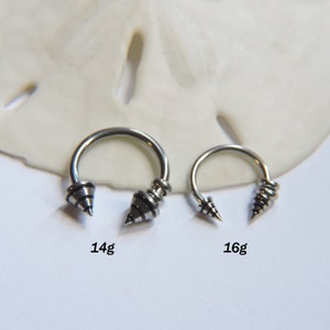 May include: Two silver circular barbell earrings with black spiral designs on the ends. The larger earring is labeled 14g and the smaller earring is labeled 16g.