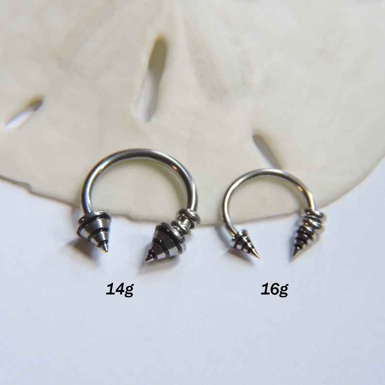 May include: Two silver circular barbell earrings with black spiral designs on the ends. The larger earring is labeled 14g and the smaller earring is labeled 16g.