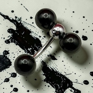Puede incluir: Un barbell para piercing corporal con tres esferas negras brillantes y una esfera central plateada. El barbell tiene una barra curva de color plateado. El fondo es blanco con salpicaduras de pintura negra.