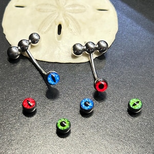 May include: A collection of body jewelry, including two curved barbells with silver-colored balls and eye-shaped gems in blue and red. Additional eye-shaped gems in red, blue, and green are also present. The jewelry is displayed on a dark surface.