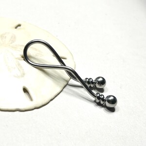 May include: A silver-toned metal body jewelry piece featuring a curved, twisted bar design that forms an elongated loop. The ends of the bar are finished with small, polished spherical beads, each secured by a decorative coiled metal detail.