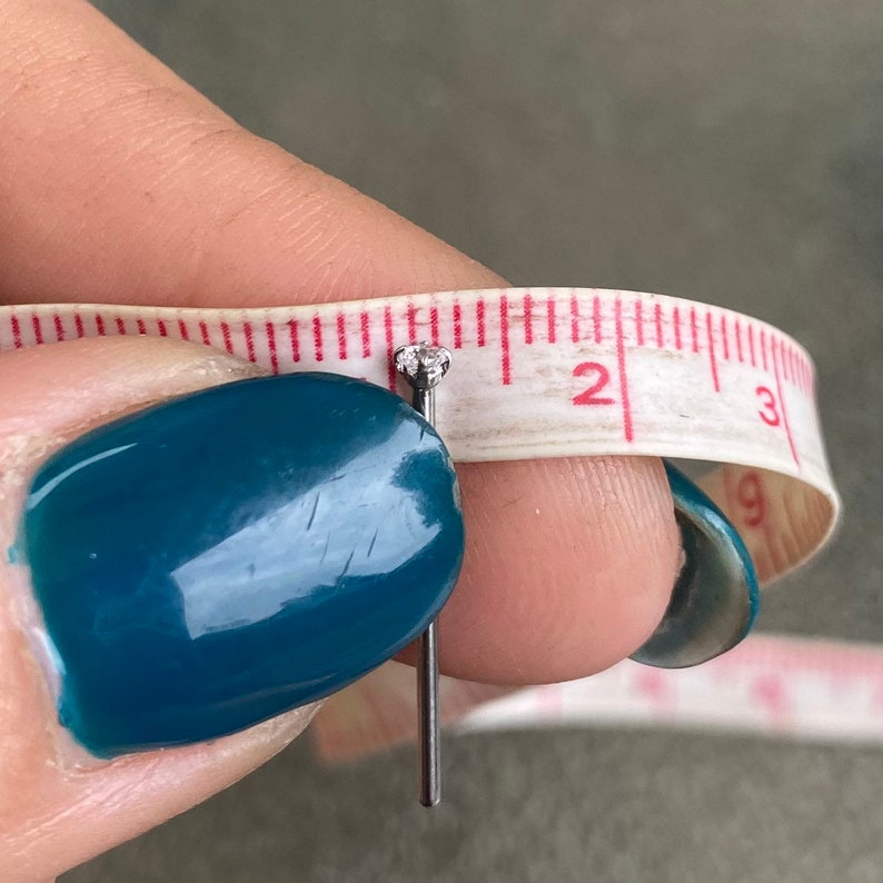 May include: A close-up of a silver nose stud with a small clear gemstone. The stud is measured against a tape measure, showing it is just under 2 inches long.