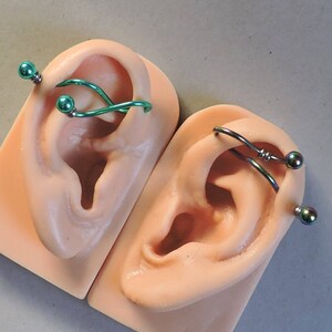 May include: Two industrial ear piercings displayed on beige silicone ear models. One piercing features a metallic green wavy bar with matching spherical ends. The other piercing consists of a dark iridescent wavy bar with two small rings and spherical ends.
