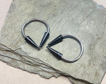 May include: Two silver-colored, U-shaped earrings with black, cone-shaped ends. The earrings are displayed on a gray, layered stone surface. The jewelry is designed for body piercing.