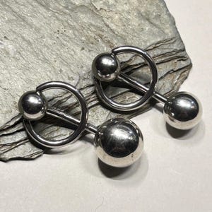 May include: A pair of silver nipple rings. Each piece has a large, spherical ball at one end, a smaller ball, and a circular design. The jewellery is presented on a grey, textured surface.