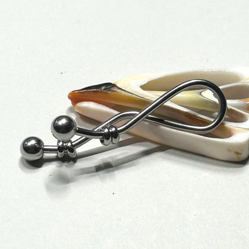 May include: A silver-toned metal body jewelry piece featuring a curved bar with two polished spherical ends and a looped, infinity-shaped design. The jewelry is positioned against a smooth, cream and orange-toned seashell with a hollow center.