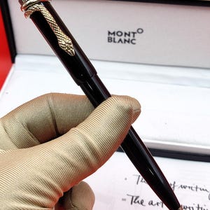 Pode incluir: Caneta Montblanc preta com um design de cobra dourada enrolada na parte superior. A caneta tem uma ponta prateada e é apresentada em uma caixa branca com o logotipo Montblanc.