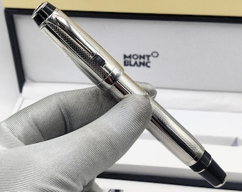 May include: A silver-toned Montblanc fountain pen with a textured, cross-hatched metal finish, featuring a black cap top and a black end piece. The pen includes a silver clip and is presented in a branded box with the Montblanc logo visible.