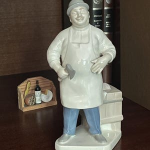 Może przedstawiać: Porcelanowa figurka rzeźnika w białym fartuchu, jasnoszarym kapeluszu i niebieskich spodniach. Postać trzyma w prawej dłoni mały szary tasak i stoi obok białego bloku. Obok znajduje się mała drewniana skrzynka z artykułami spożywczymi.