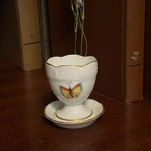 May include: A white porcelain egg cup with a matching saucer, featuring a scalloped rim and a gold-trimmed edge. The cup is decorated with a colourful butterfly illustration in shades of yellow, orange, and green, and has a textured basket-weave pattern.