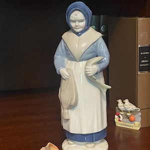 Może przedstawiać: Porcelanowa figurka przedstawiająca osobę w niebieskim czepku, jasnoniebieskiej bluzce z długim rękawem i białym fartuchu. Postać trzyma rybę w lewej dłoni i worek w prawej, stojąc na okrągłej podstawie z muszlą umieszczoną z przodu.