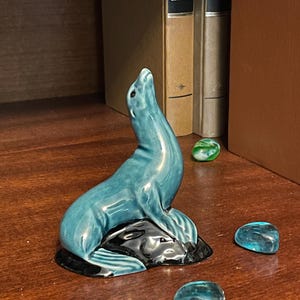 Può includere: Una statuetta in ceramica lucida raffigurante un leone marino dal corpo color verde acqua, posizionato con la testa rivolta verso l'alto. Il leone marino poggia su una base nera simile a una roccia. Tre piccole pietre di vetro blu e verde.