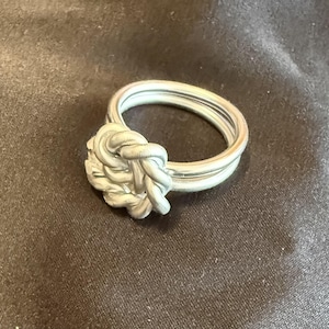 Op de afbeelding: Een zilverkleurige ring met een opvallend, ingewikkeld knoopontwerp aan de voorzijde. De ringband is opgebouwd uit meerdere dunne, gladde, draadachtige strengen die samenkomen in een centraal, getextureerd, gevlochten knoopelement.