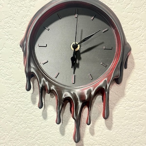 May include: A round wall clock with a matte black face and simple black hour markers. The frame features a distinctive melting effect, with black and deep red drips cascading down the sides and bottom, creating a fluid, irregular silhouette.