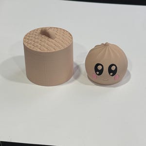 May include: A light tan 3D printed cylindrical bamboo steamer basket with a textured lid alongside a matching round dumpling figurine featuring a smooth surface, large black eyes, a small mouth, and pink circular cheeks on its front side.