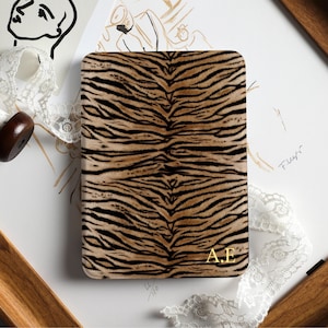 Puede incluir: Una funda rectangular para lector electrónico con un patrón de rayas de tigre en marrón y negro. La esquina inferior derecha de la funda muestra las iniciales A.E. en color dorado. La funda tiene esquinas redondeadas y una superficie lisa.