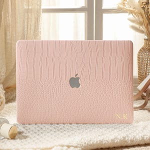 Może przedstawiać: Jasnoróżowe twarde etui na laptopa z teksturowanym wzorem skóry krokodyla. Etui posiada centralne wycięcie na logo Apple oraz złote inicjały N.K. nadrukowane w prawym dolnym rogu prostokątnej pokrywy.