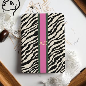 May include: A rectangular notebook with a zebra print cover, a pink vertical stripe, and the gold letters "A.E." The notebook is surrounded by art supplies and a wooden frame.