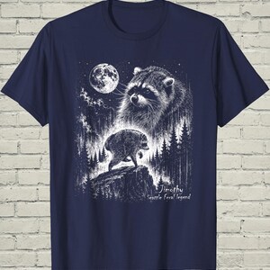 May include: A navy blue t-shirt featuring a white graphic illustration of a raccoon in a forest at night under a full moon. The design includes a large raccoon face above a smaller raccoon standing on a rock, with the text Jimothy Seattle Feral legend.