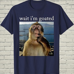 May include: A navy blue short-sleeve t-shirt featuring a central graphic of a brown monkey with its hand near its chin, set against a coastal background. Above the image, the white text reads, wait i'm goated, in a simple serif font.