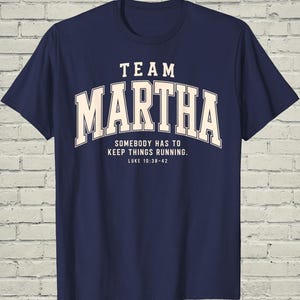 May include: A navy blue short-sleeve t-shirt featuring the text "TEAM MARTHA" in large cream-colored block letters, followed by the text "SOMEBODY HAS TO KEEP THINGS RUNNING." and the reference "LUKE 10:38-42" printed in a smaller cream font.