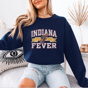 May include: A navy blue crewneck sweatshirt featuring the text "INDIANA FEVER" in a distressed white font with red outlines. Between the words is a yellow banner graphic with a stylized letter "F" that includes a basketball and flame detail.