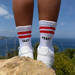 Könnte beinhalten: Weiße Crew-Socken mit zwei horizontalen roten Streifen am oberen Rand und dem schwarzen Schriftzug "EASY" auf der linken sowie "PEASY" auf der rechten Socke, kombiniert mit weißen Sportschuhen, die das Nike-Logo auf der Ferse tragen.