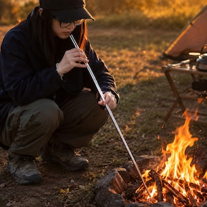 Peut inclure: Un long tube de soufflet de feu en métal argenté, fin et télescopique, utilisé pour diriger le flux d'air vers un feu de camp. L'outil est tenu par les deux extrémités, avec une pointe placée près de la base du bois pour attiser les flammes.