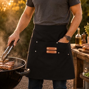May include: Black apron with two pockets and leather accents. The apron has a simple design with metal rivets and is being worn while grilling. The person is using tongs to handle food on a charcoal grill.