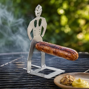 May include: A stainless steel grilling tool shaped like a stylized human figure, holding a cooked sausage. The figure has cut-out features and stands on a square base. The sausage is brown and appears freshly grilled, with steam rising from the grill.