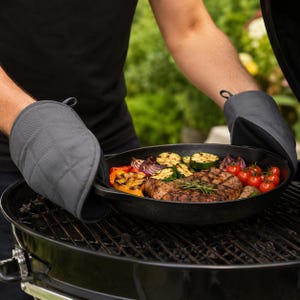 Peut inclure: Une paire de gants de cuisine matelassés gris foncé tenant une poêle en fonte noire contenant un steak grillé, des tomates cerises, des courgettes tranchées, de l'oignon rouge et des poivrons, avec un brin de romarin frais sur la viande.