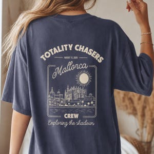 May include: A navy blue t-shirt featuring a cream-colored graphic on the back. The design includes the text "TOTALITY CHASERS", "AUGUST 12, 2026", "Mallorca", "CREW", and "Exploring the shadows" alongside an illustration of a sun and city skyline.